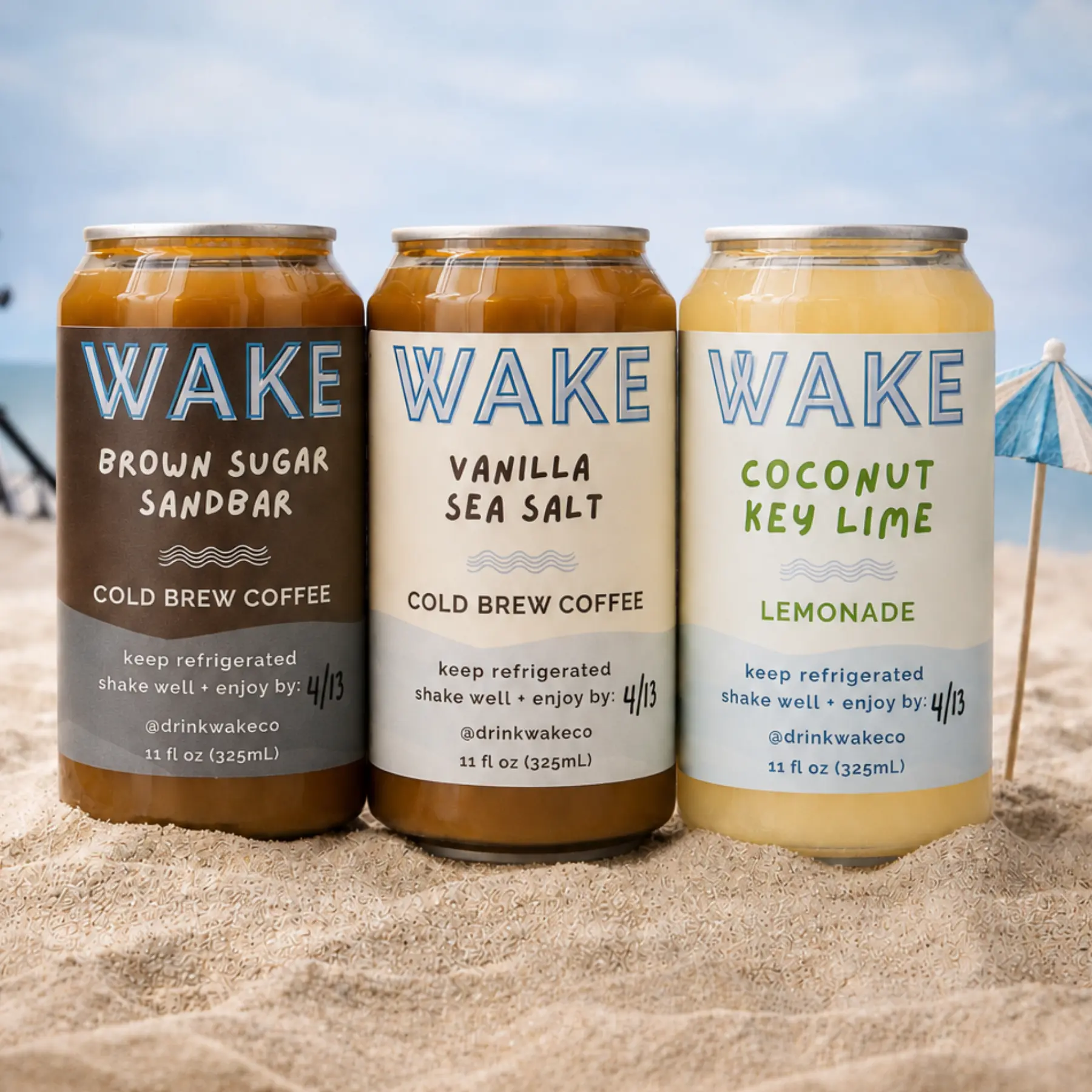 Three WAKE cans — Brown Sugar Sandbar, Vanilla Sea Salt and Coconut Key Lime — standing in the sand at the beach